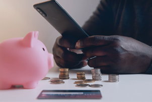 An image of a person sitting at a table with a piggy bank, credit card, and stacks of coins on it, while scrolling on their phone.