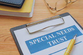 Image of a paper on a clip board that says "SPECIAL NEEDS TRUST" surrounded by a pen, glasses, and books.